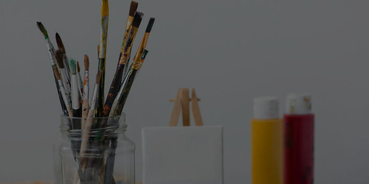 A set of paintbrushes in a clear glass jar, with two paint tubes and a small easel visible against a plain gray background.