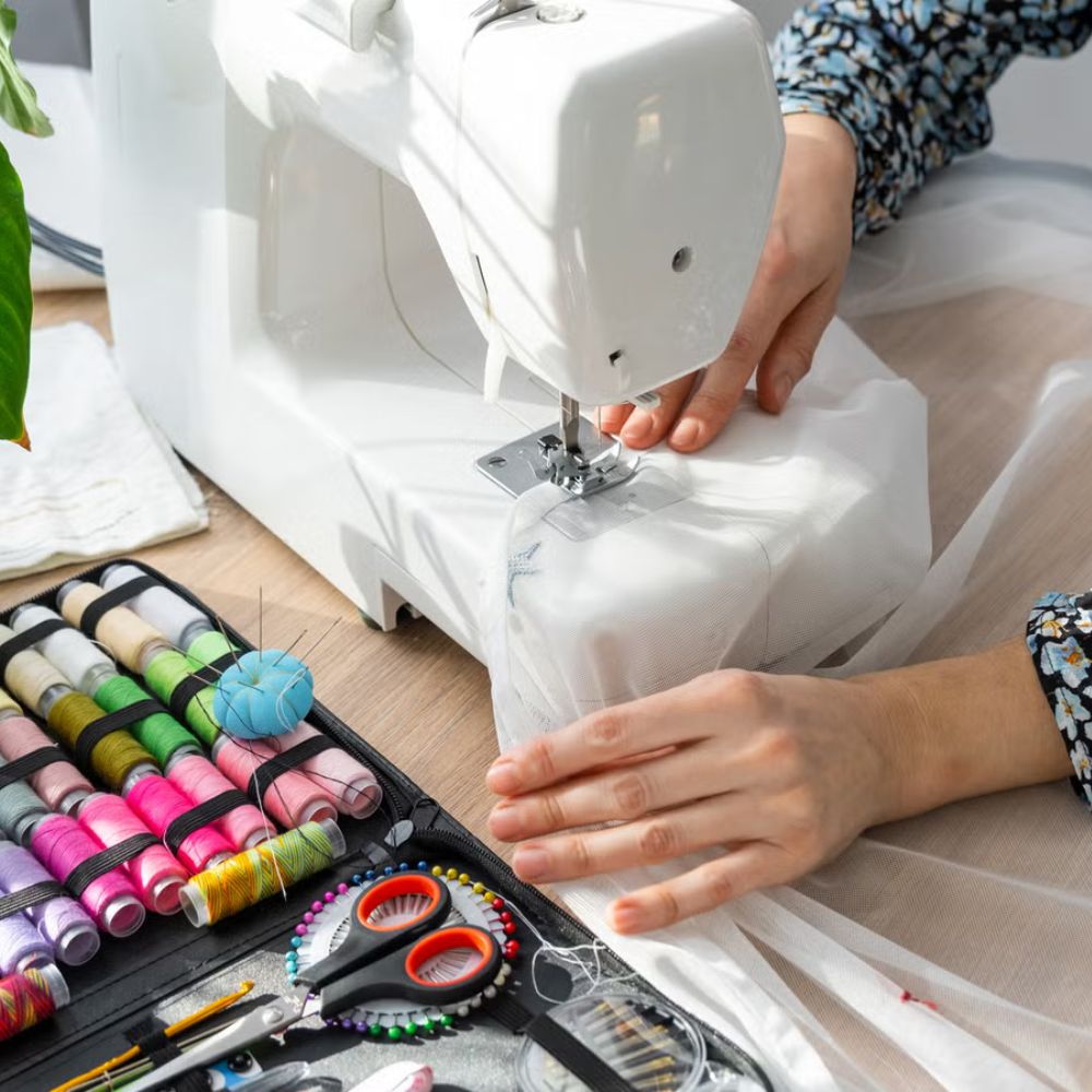 Person using a sewing machine with a tray of colorful thread and scissors on a table.