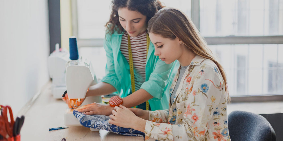 Two women working at a sewing table in a bright room. One woman in a teal jacket is operating a white sewing machine, while the other in a floral blouse holds a piece of blue patterned fabric.