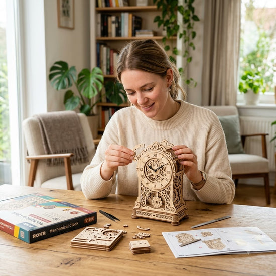 A woman sits at a wooden table in a bright living room, assembling a decorative mechanical clock. The kit includes the assembled clock, loose wooden pieces, a colorful instruction box, and printed instructions on the table.