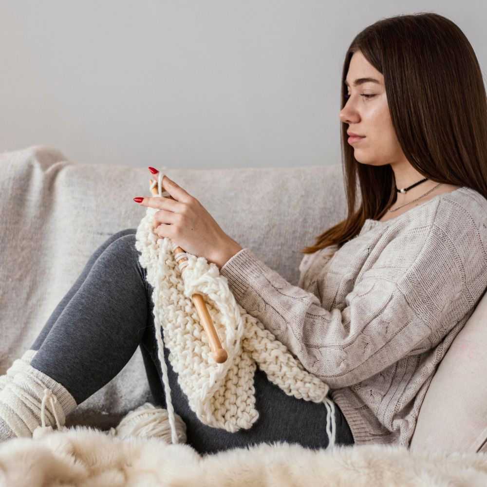 Alt Text: Person knitting chunky cream yarn project relaxing comfortably on sofa